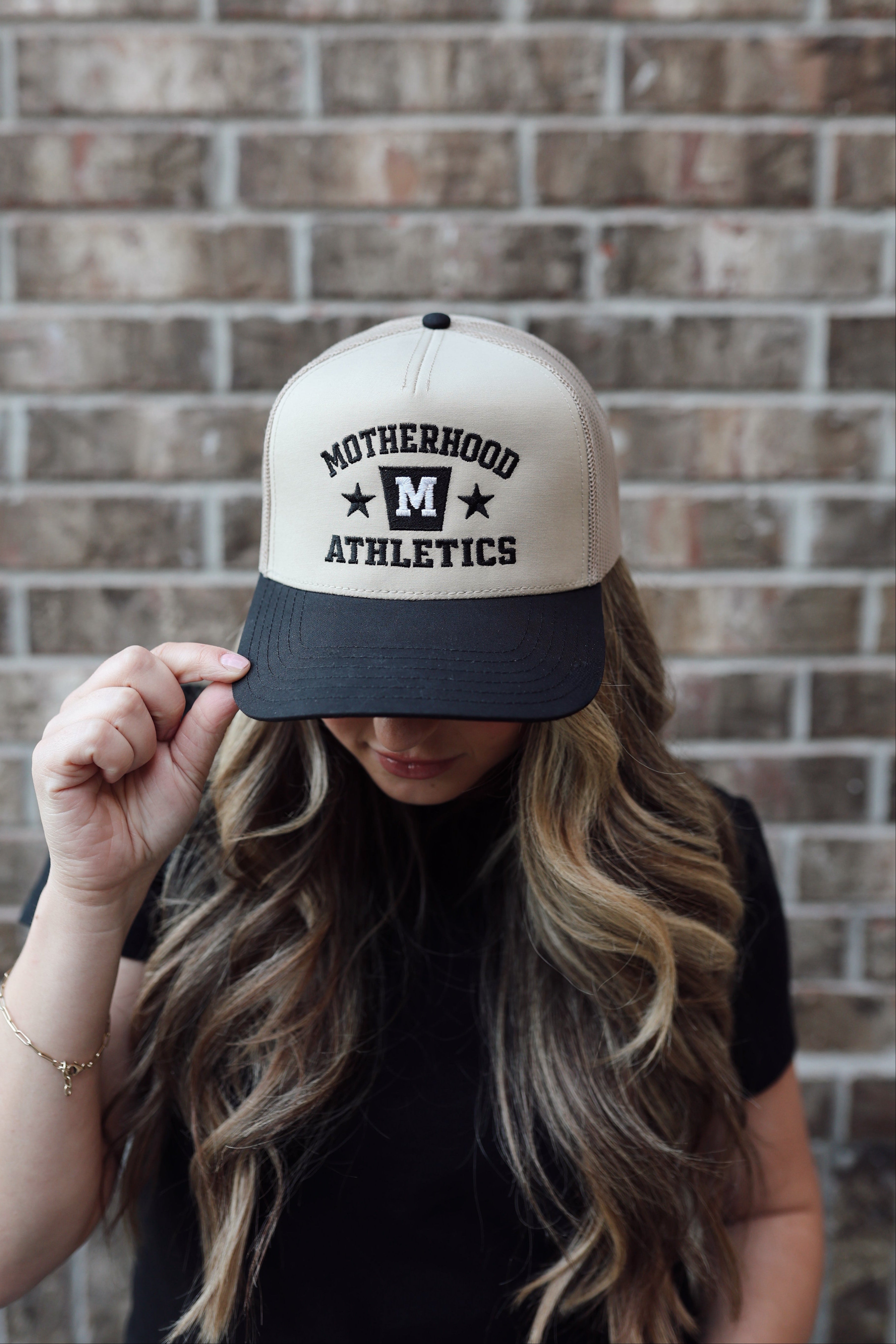 mom wearing a cap with 'Motherhood Athletics' text against a brick wall. 