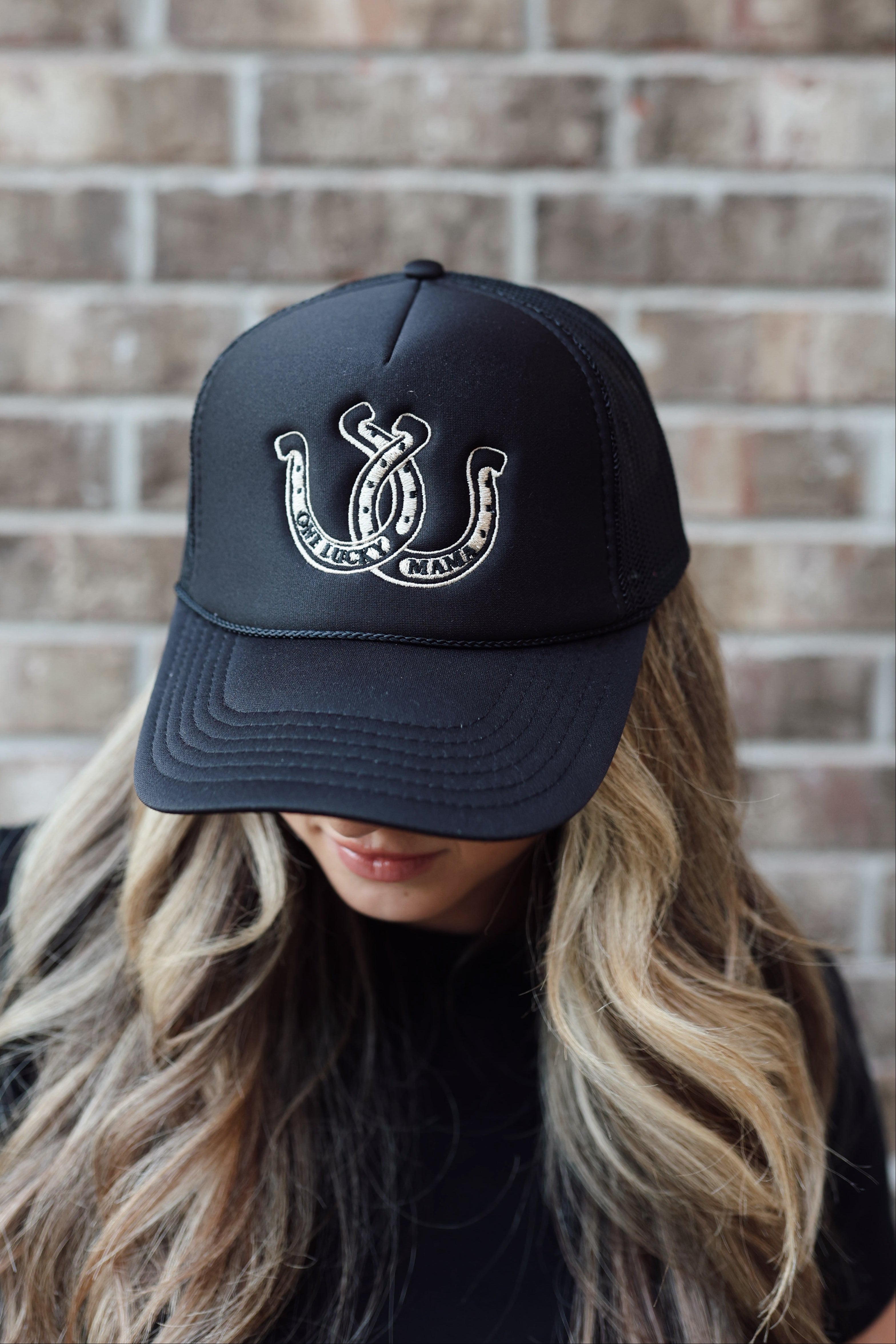 Mom wearing a mom hat that is all black with a western embroidered design of a horseshoes on it that says "One Lucky Mama". Cute mom trucker hat